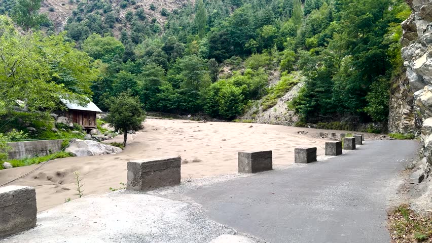 Muddy floodwater surges beside a mountain road through a forested valley in Kashmir