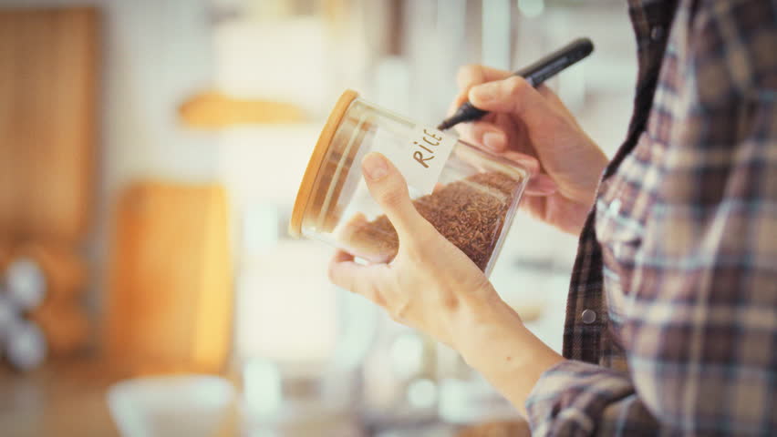 In a home kitchen, a girl labels a clear glass jar rice for cereal storage. The tidy act showcases her habit of organizing the kitchen, ensuring proper food preservation and household order