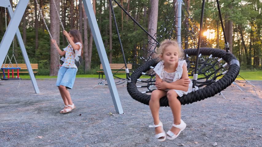 Two girls enjoy their time at a park in the evening. One girl swings on a swing while another sits on a nest swing. Trees surround the play area