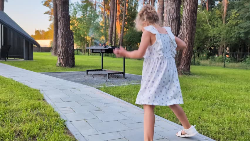 A child walks carefully on a stone path in a park surrounded by tall trees. A grill is visible in the background as the sun sets