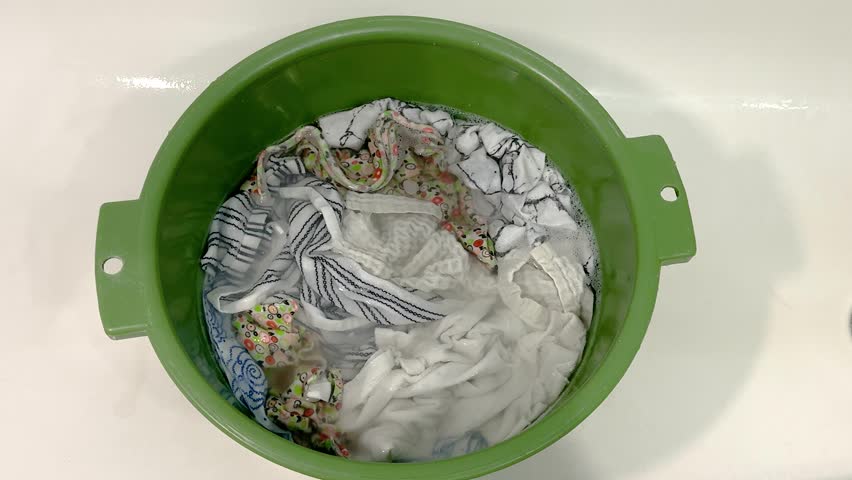 White clothes soaking in soapy water inside a green plastic basin. Hand laundry, household chores, cleaning routine.