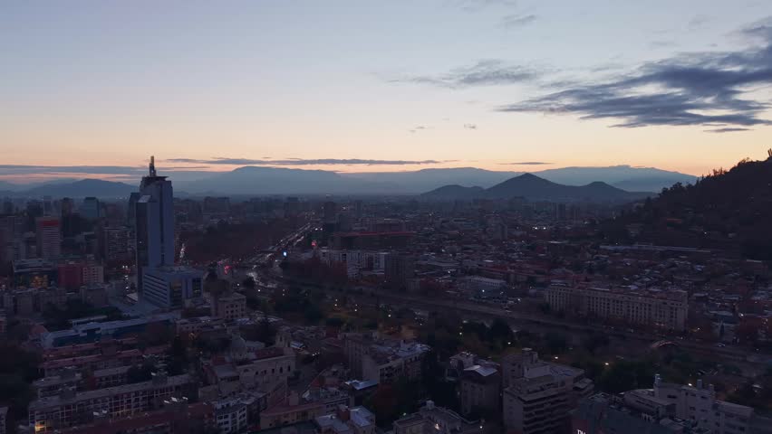 Advancing flight over a wide city skyline with a highway and river winding below toward distant hills and mountains at dusk. Santiago, Chile
