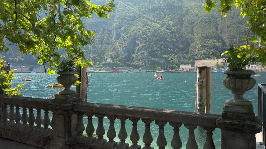 Beautiful waterfront terrace overlooking turquoise Lake Garda in Riva del Garda, Italy, framed by a historic stone balustrade, decorative urns and leafy trees. Small boats and water activities animate the lake while steep mountains rise behind the picturesque resort town. Scenic summer tourism and European vacation destination in Trentino Alto Adige, ideal for travel, leisure, nature and relaxation concepts.