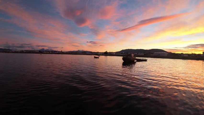 A silhouetted fishing boat and small canoes rest on calm water beneath vivid pink and orange clouds near Puerto Natales Chile, FPV aerial