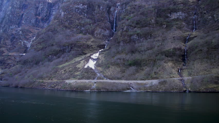Steep fjord cliffs with narrow waterfalls and snow patches above calm water in Norway