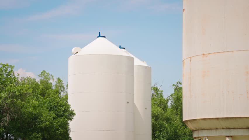 Static shot of white grain silos on a prairie farm with green trees swaying in the wind beneath a bright blue summer sky