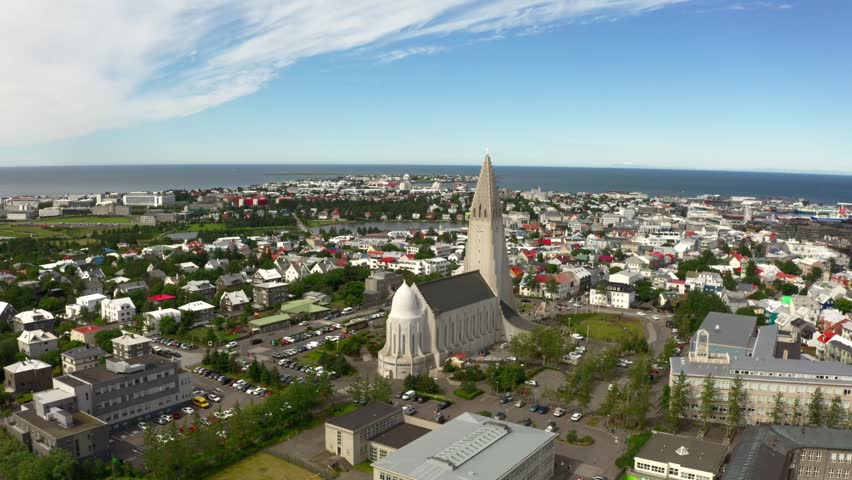 Aerial view of Reykjavik, Iceland, on a sunny day. The cityscape features the iconic Hallgrímskirkja church, colorful buildings, and the ocean in the background.