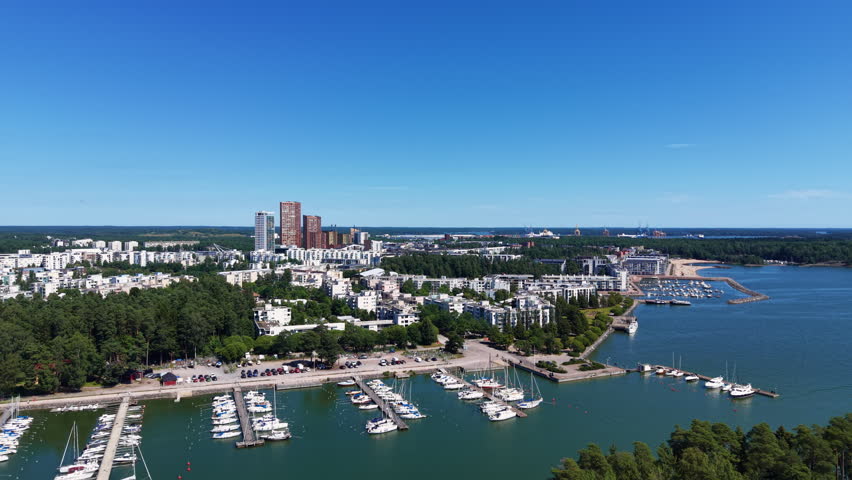 Vuosaari district in Helsinki, Finland, showing a marina with sailboats, residential buildings amid green trees, and the Baltic Sea under a clear blue sky