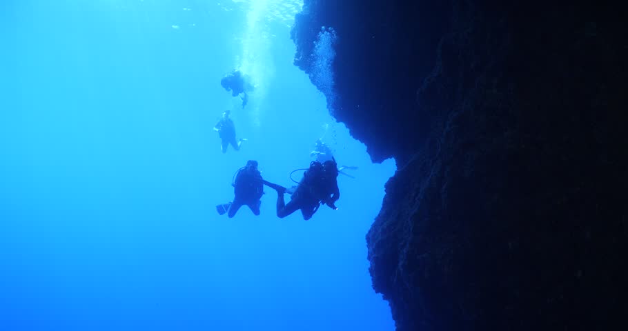  scuba divers exploring around a reef underwater deep blue water big rocks and bubbles ocean scenery 
