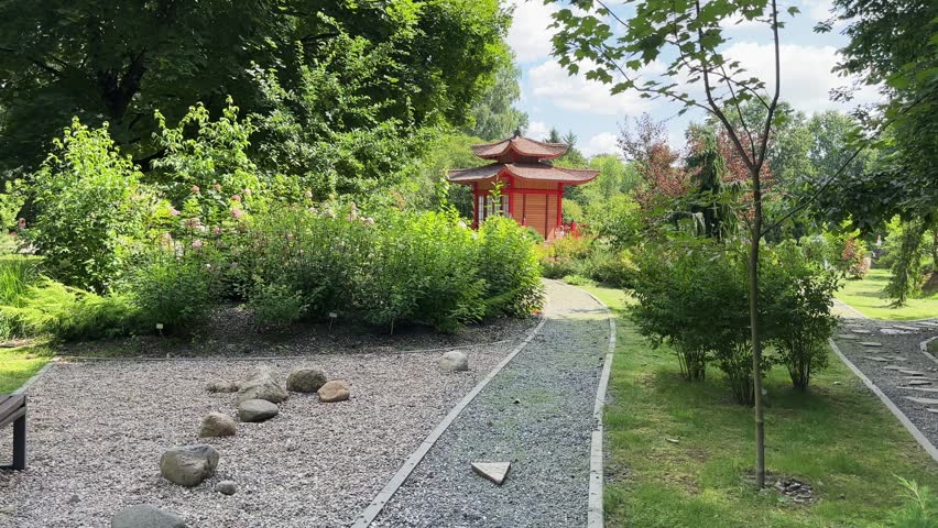 Scenic Japanese garden path leads past green shrubs toward red wooden pagoda. Summer sky with light clouds hangs over oriental pavilion and stone walkway. Landscape framing captures Asian design.