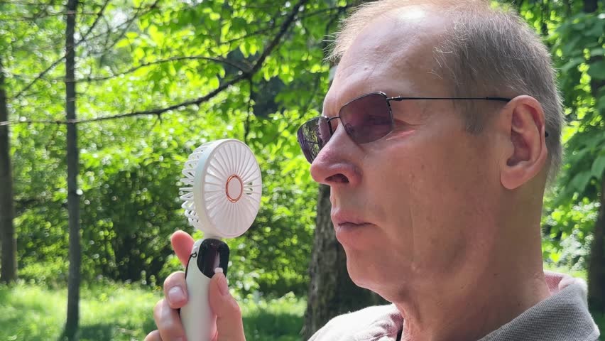 Mature man wearing UV protection sunglasses using portable mini fan in sunny park. Complete sun and heat protection concept during hot summer weather.
