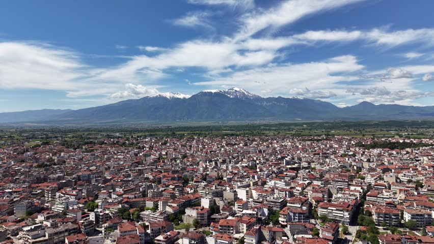 Forward aerial drone flight above the dense urban landscape of Katerini toward the snow-capped Mount Olympus mountain range under a blue sky with dramatic white clouds.