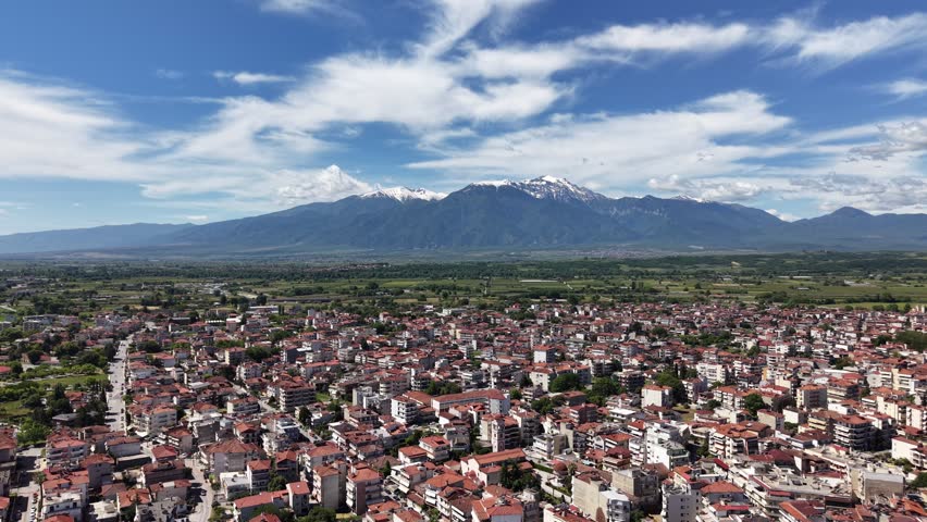 Forward aerial drone flight above the dense urban landscape of Katerini toward the snow-capped Mount Olympus mountain range under a blue sky with dramatic white clouds.