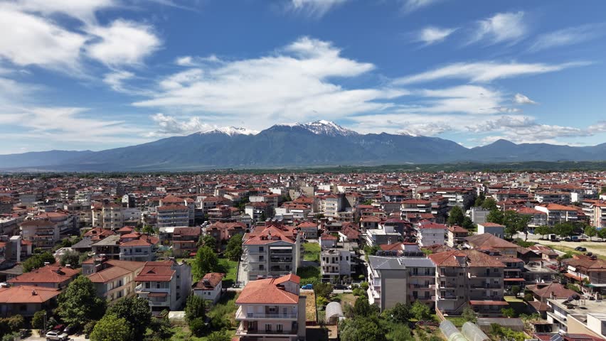 Descending aerial drone view above the dense urban landscape of Katerini, with red-roofed buildings and the snow-capped Mount Olympus range visible beneath dramatic