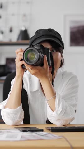 A woman in her thirties tests a camera while dressed in a white shirt and black cap at a modern workspace with photo prints and bright lighting around her.