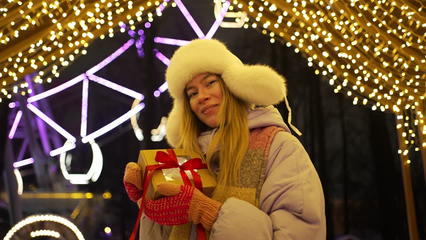 Happy woman wearing winter clothes holding a wrapped present and smiling at an amusement park. Bright festive lights and a ferris wheel decorate the cheerful night atmosphere