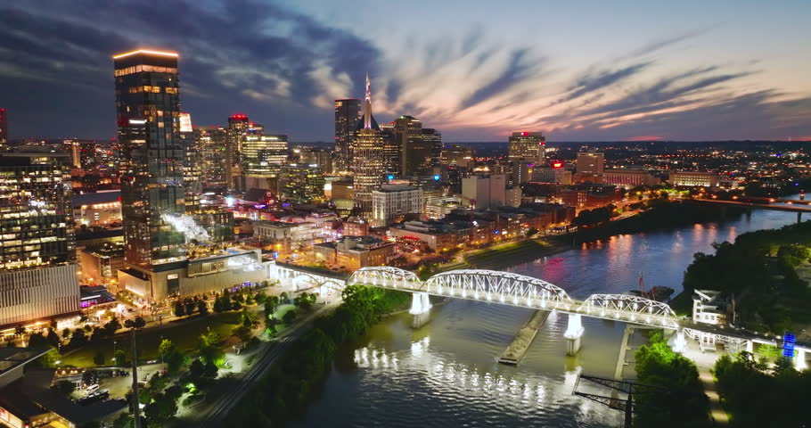 Aerial panorama of downtown Nashville, Tennessee at night with modern skyscrapers, river waterfront and city bridge illuminated by city lights.