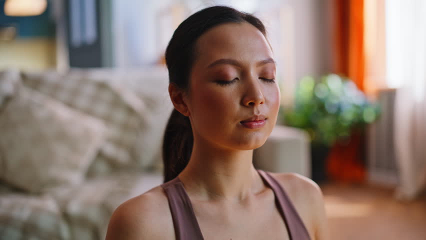 Calm asian woman finishing breathing meditation home living room, opening eyes smiling at camera. Closeup face portrait of relaxed mindful girl in sportswear, peaceful wellness mindfulness lifestyle.