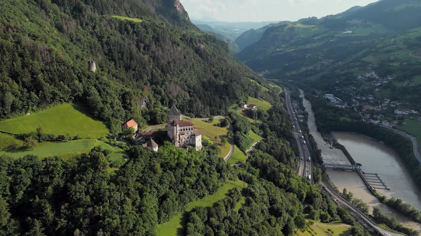 Trostburg Castle on forested hill with river and highway running through alpine valley, South Tyrol Dolomites, aerial view