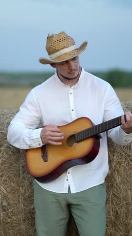 Young man in straw cowboy hat plays acoustic guitar leaning on hay bale in rural field under cloudy sky. Authentic country lifestyle and folk music scene.