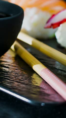 A close view of sushi arranged on a black plate with chopsticks placed beside it. The setting includes various sushi types and a small bowl likely for sauce.