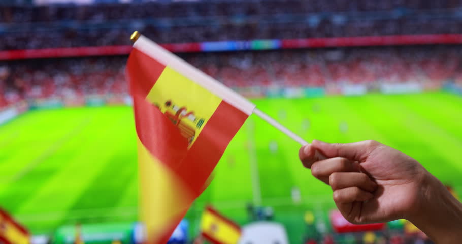 Hand holding a spain flag in a soccer stadium with blurred field background