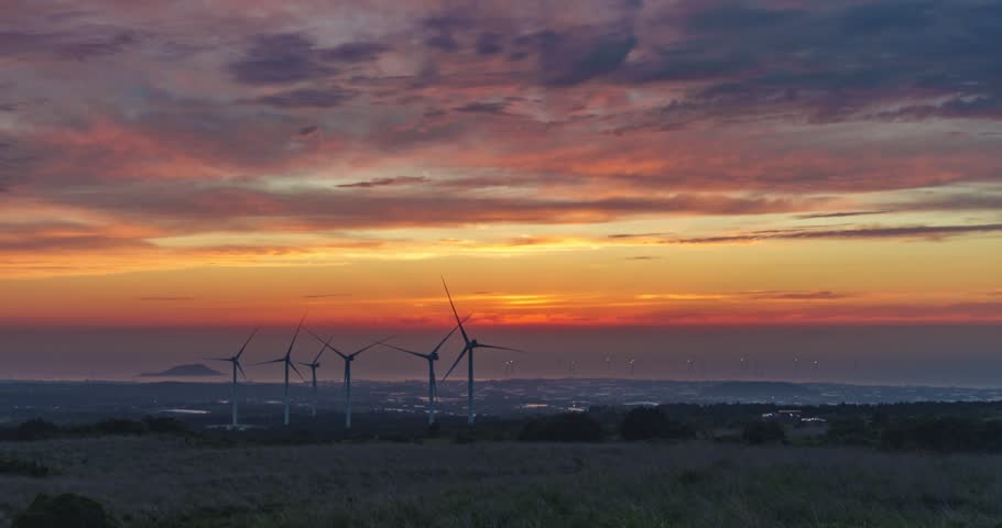 Silhouetted wind turbines stand against vivid orange sunset sky above Jeju coastline with distant island and city lights - time lapse