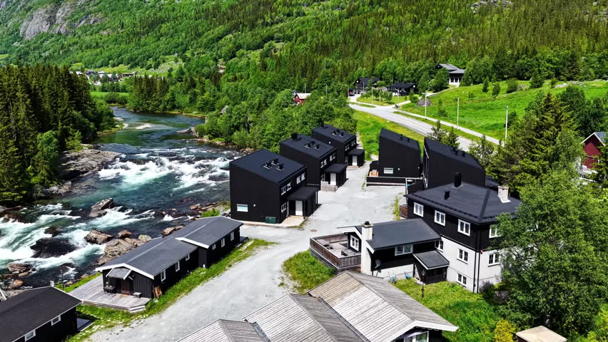 Black Riverside Cabins Sit Beside Rushing Rapids Along the Road From Flåm to Oslo, Norway - Aerial Drone Shot