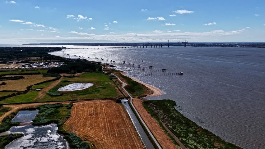 Aerial view over farmland and carrelet fishing huts along Loire Estuary shoreline with Saint-Nazaire Bridge in distance France