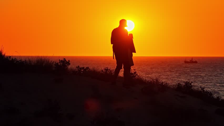 Silhouette of man standing on coastal cliff watching vivid orange sunset over ocean with distant boat sailing near horizon