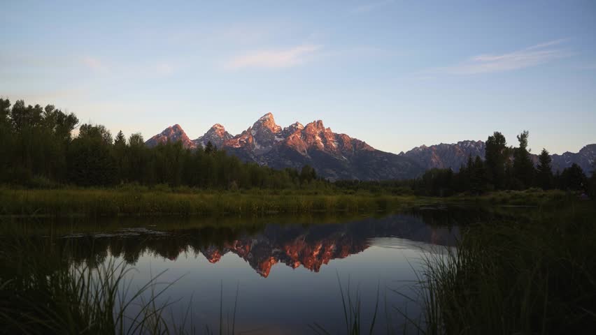 Summer sunrise illuminates the towering peaks of Grand Teton National Park as colorful light reflects across the calm Snake River at Schwabacher Landing, surrounded by lush green trees and brush.
