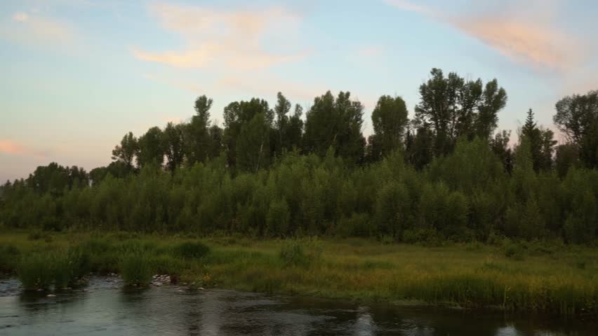 Panning right revealing a sunrise illuminates the towering peaks of Grand Teton National Park as colorful light reflects across the Snake River at Schwabacher Landing, surrounded by green brush.