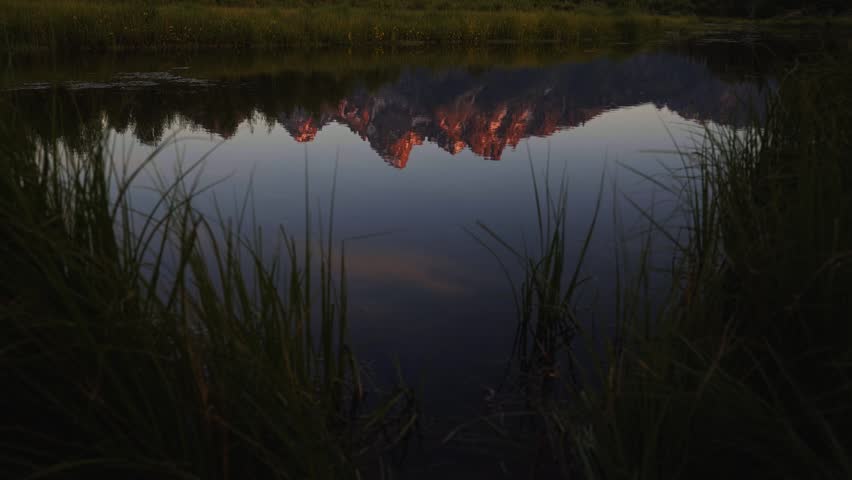 Tilting up revealing a sunrise illuminates the towering peaks of Grand Teton National Park as colorful light reflects across the Snake River at Schwabacher Landing, surrounded by green brush.