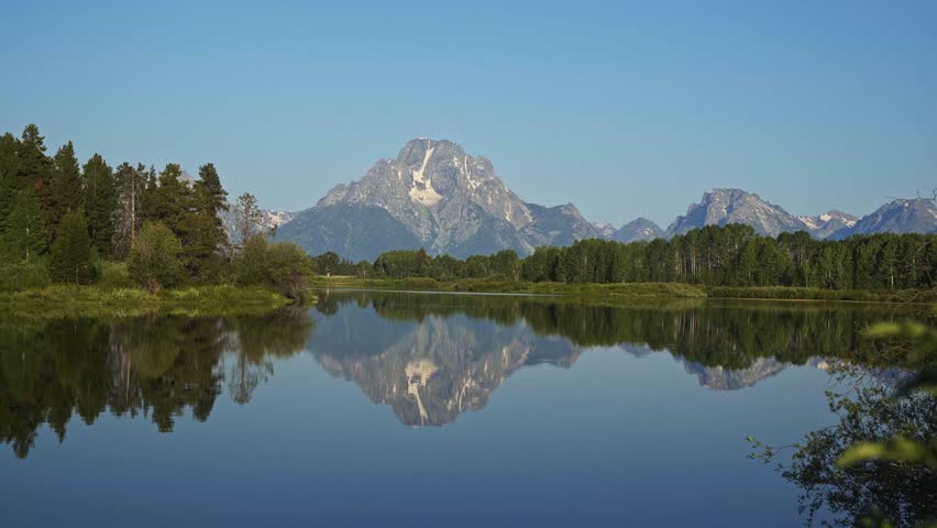 Mount Moran rises above the calm waters of Oxbow Bend in Grand Teton National Park, with its striking reflection mirrored below and lush summer greenery framing the peaceful Wyoming landscape.