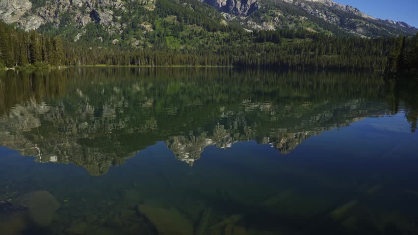 Sunny summer view of Taggart Lake in Grand Teton National Park with crystal-clear waters reflecting the towering Teton Mountains, surrounded by lush pine forests and peaceful alpine scenery in Wyoming