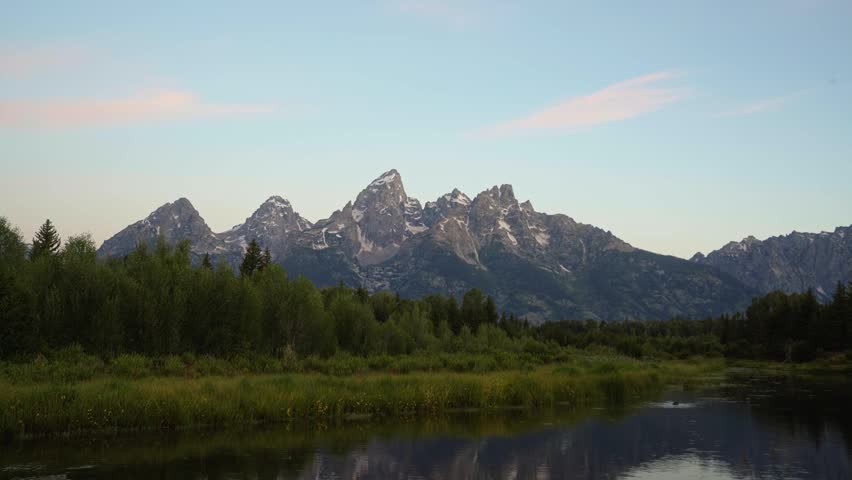 Tilting down revealing a sunrise illuminates the towering peaks of Grand Teton National Park as colorful light reflects across the Snake River at Schwabacher Landing, surrounded by green brush.