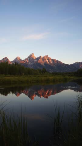Vertical shot of a summer sunrise illuminates the towering peaks of Grand Teton National Park as colorful light reflects across the Snake River at Schwabacher Landing, surrounded by lush green brush.