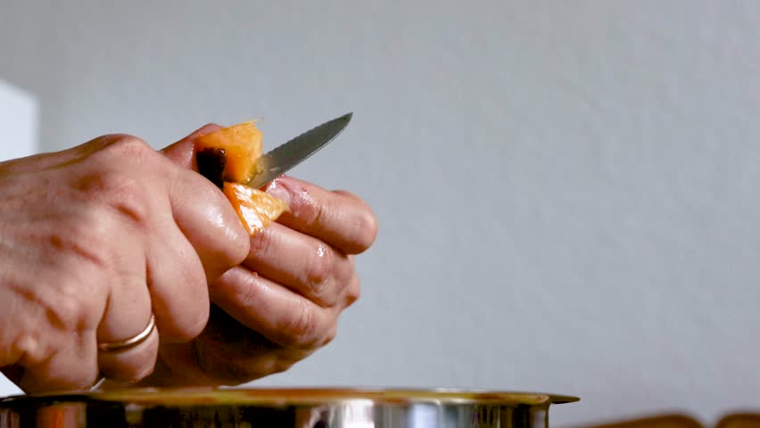 Closeup of a woman hands cutting and slicing a very ripe, juicy peach with a small knife over a metal mixing bowl. 
