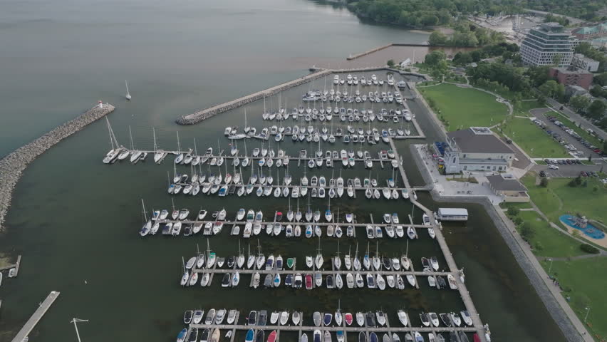 Numerous sailboats sit docked in a protected marina basin next to green waterfront parkland in Oakville, Ontario. A rocky breakwater separates the calm harbour from open lake water.
