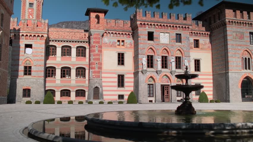 Front facade of Castello Ceconi stands in sharp focus against a vivid blue sky, its reflection visible in a softly blurred fountain, northern Italy.