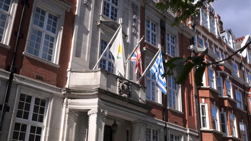 The Hellenic Centre in Marylebone is framed from below as Greek, British and Cypriot flags move gently against the sunlit facade.