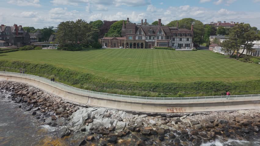 Newport Rhode Island Cliff Walk promenade, grand Tudor-style mansion, manicured lawn, rocky coastline, ocean waves, coastal path, historic estate, New England shoreline. Aerial drone view.