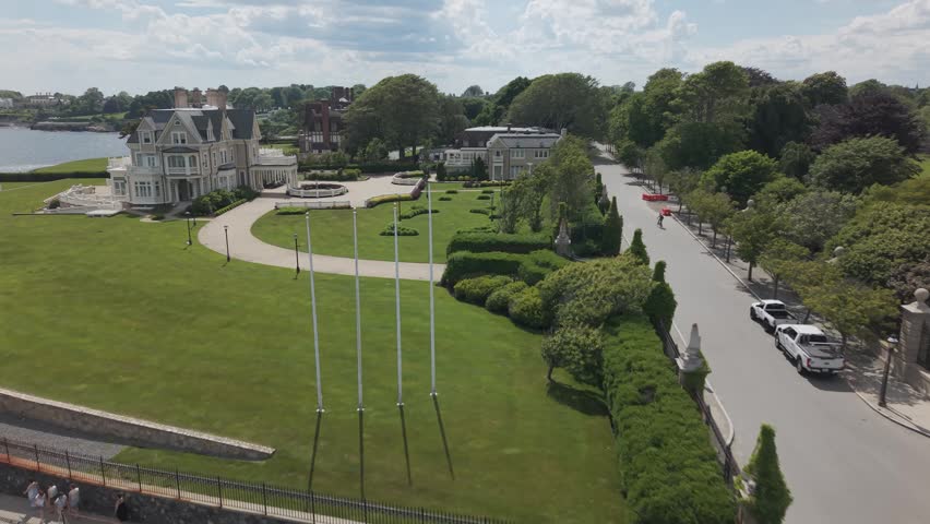 Newport Rhode Island Gilded Age mansion, circular driveway, flagpoles, manicured gardens, waterfront estate, tree-lined street, historic architecture, New England coast. Aerial drone approaching shot