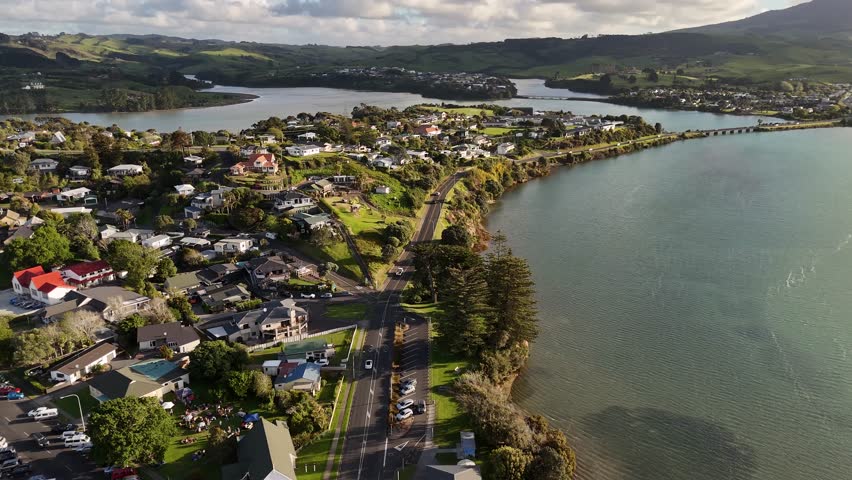 Winding coastal road passes waterfront homes and parked cars near estuary shoreline at Raglan, New Zealand, distant bridge and green hills rise beyond calm blue water. Aerial drone wide shot