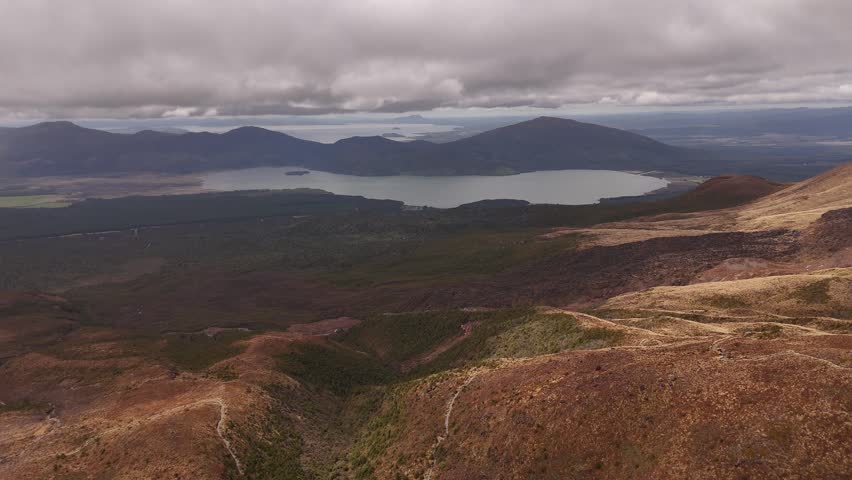 Winding hiking trails cross golden volcanic slopes above distant lake at Tongariro, North Island, New Zealand, layered mountain ranges stretch beneath heavy grey clouds. Aerial drone wide shot