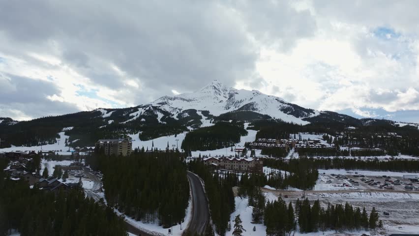 Trucking left aerial drone shot of a winter landscape of a mountain resort in Montana with Lone Peak rising in the background, surrounded by pine forests, homes, and snow-covered terrain.