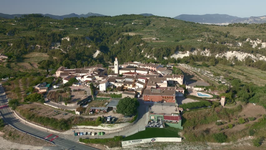 Aerial perspective slowly orbiting the picturesque village of Benillup in Alicante, Spain, showcasing its charming church, traditional homes, and surrounding green mountains - stock photo