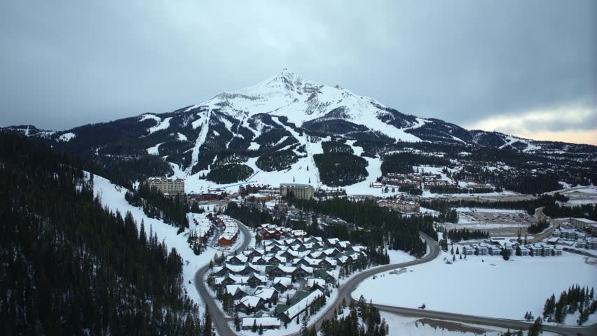 Dolly in aerial hyperlapse of a Montana mountain resort landscape with Lone Peak in the distance, featuring snowy forests, buildings, moving vehicles, and active ski infrastructure on a March day.