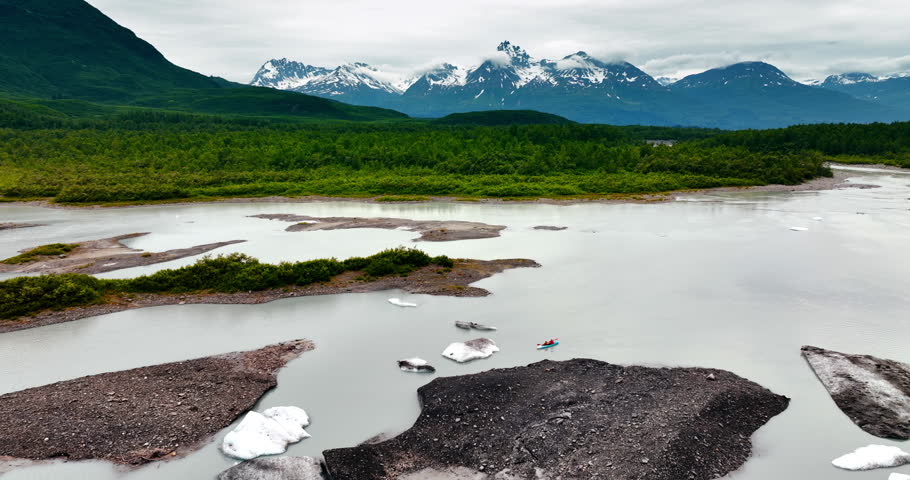 Two people row in canoe by the river with large pieces of ice in it. Amazing green waterfront and spectacular mountains with snow on the slopes at backdrop. Alaska.