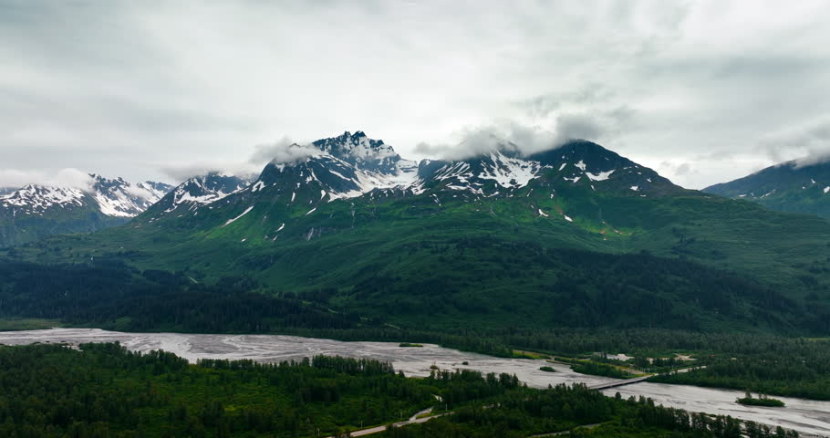 Beautiful verdant mountains with some snow on the tops. Grey clouds cover the peaks of the rocks. River flows at the mountain foot. Alaska aerial.